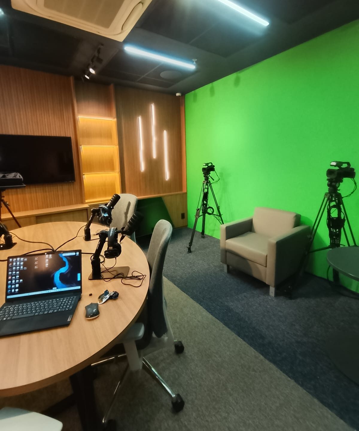 Estúdio do CFM com a bancada de podcast, o cenário em madeira com luz de LED e, ao lado, o fundo verde de chroma key com câmeras