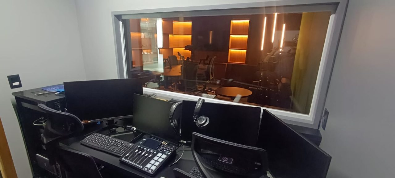 Sala de controle do estúdio do CFM: mesa de corte e monitores diante do visor de vidro para o estúdio de podcast